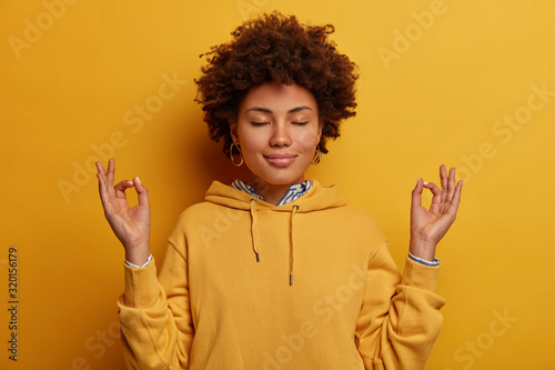 Portrait of ethnic woman stays calm, meditates and practices yoga, keeps hands in zen gesture, closes eyes, relaxes after hard working day, wears yellow sweatshirt, poses indoor, unites with nature