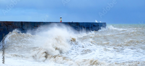 A harbour wall with a rough sea crashing against it and a lighthouse in the background, Newhaven harbour, Sussex, England