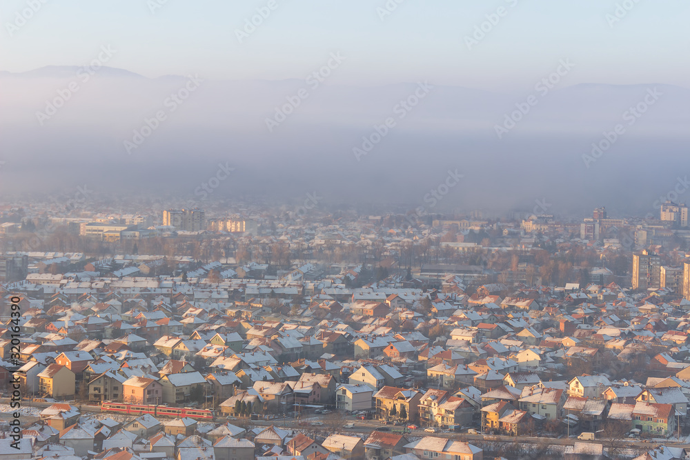 Red train passing in front of misty cityscape with buildings lighten by golden sunrise and rooftops covered by first snow