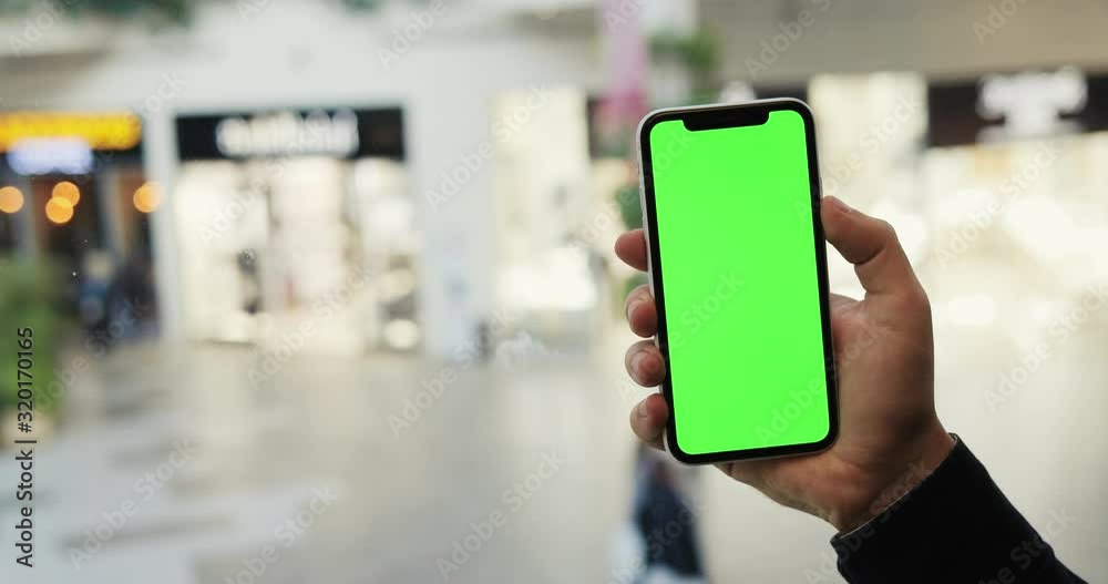 Close up of the hand of Caucasian man holding vertical black smartphone with green screen and tapping on it in the elevator of shopping mall. Chroma key. Inside.