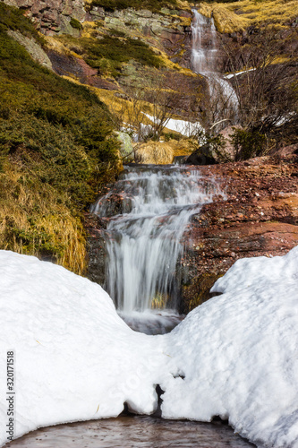 Low perspective view of large  scenic  beautiful waterfall water falling from red rocky cliff under the snow