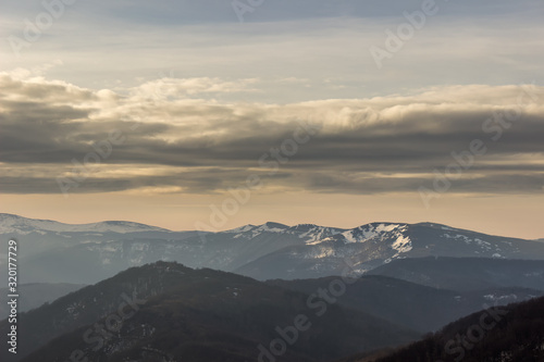 Moody, dramatic sky above pointy, distant, impressive Kom summit in Bulgaria covered by snow and misty, foreground mountain layers © Nikola
