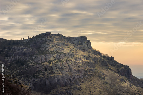 Rocky, sunlit peak on Old mountain covered by yellow grass and moss under a colorful, cloudy sky