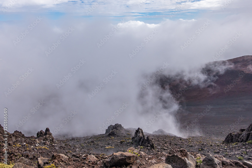 Haleakala Volcano, Maui, Hawaii, USA. - January 13, 2020: Whtie fog lingers in crater. Brown, and black rocks and yellowish ground vegetation at edge of crater. Blue patches in sky.