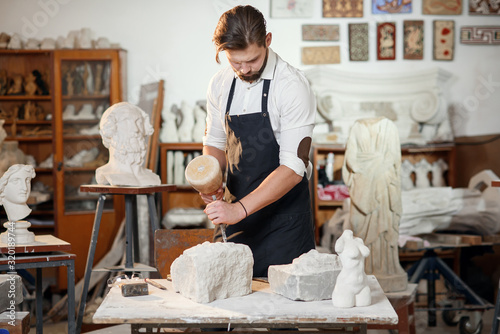 Bearded craftsman works in white stone carving with a chisel. Creative workshop with works of art. photo