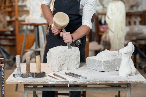 Bearded craftsman works in white stone carving with a chisel. Creative workshop with works of art.