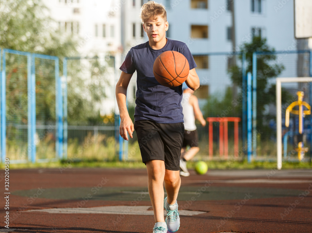 Cute smiling boy in blue t shirt plays basketball on city playground. Active teen enjoying outdoor game with orange ball. Hobby, active lifestyle, sport for kids.	