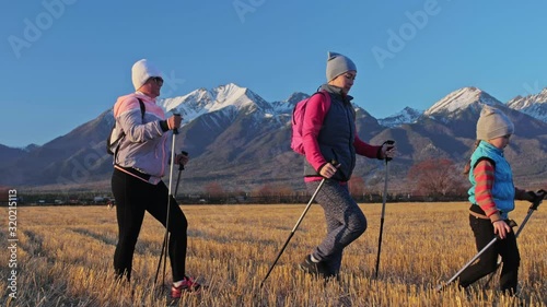 Woman Nordic walking in nature on background of mountains in field. Girls and children use trekking sticks and nordic poles, backpacks. Family travels sports. Kid is learning from mother and photo