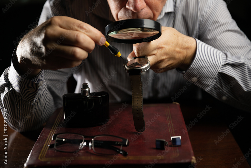 Mature man working at home as a watchmaker and jeweler composing an old clock using a screwdriver and a magnifying glass to be able to see closely