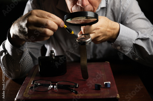 Mature man working at home as a watchmaker and jeweler composing an old clock using a screwdriver and a magnifying glass to be able to see closely
