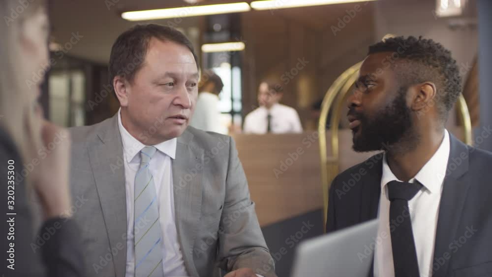 Tracking shot of middle-aged Asian businessman discussing work with male and female partners in lobby of hotel Tracking shot of middle-aged Asian businessman discussing work with male and female partners in lobby of hotel