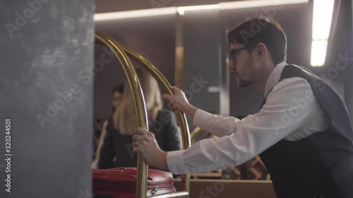 Tracking shot of serious bellhop in uniform pushing luggage cart down hotel hallway photo