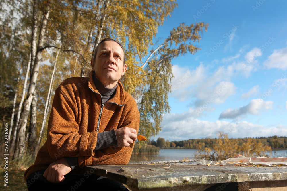 Fototapeta premium A man in an orange sweater is sitting on the street at a table from a thick board. Autumn forest