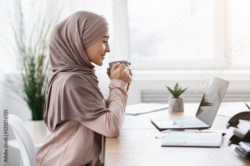 Muslim businesswoman drinking coffee at workplace while using laptop in office