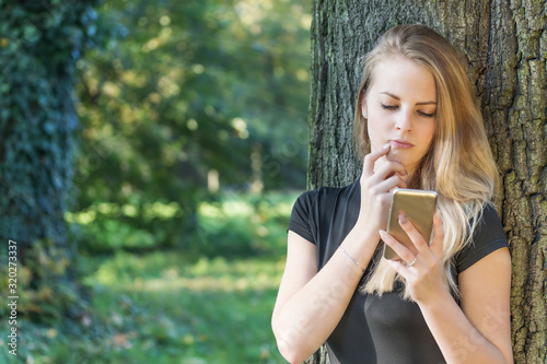 Young girl is reading message on smart phone outdoors. The girl is touching her face by right hand. All potential trademarks are removed.