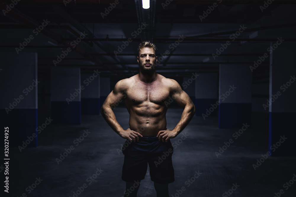 Three quarter length of topless muscular sportsman shorts standing in garage with hands on hips at night and looking at camera. Urban life concept.