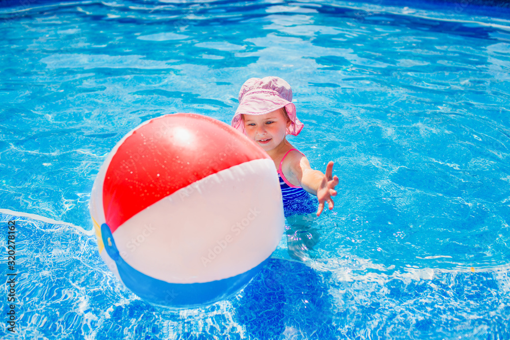 Swimming, summer vacation - lovely smiling girl in pink hat and blue swimsuit playing in blue water with inflatable multicolor ball in a pool