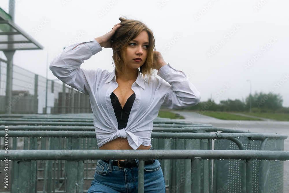 young pretty girl dressed in jeans and white shirt standing during autumn little rain on street near metal fence 