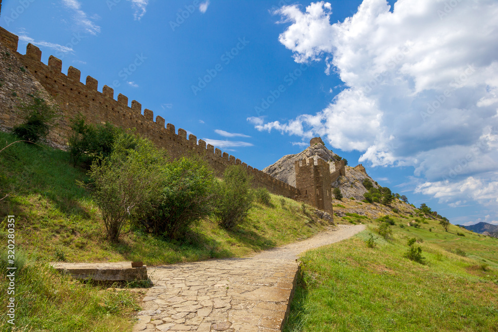 An ancient fortress on top of a mountain and path along the fortification wall in sunny day. Genoese fortress, Sudak, Crimea. Historic old building. Monument of architecture. 