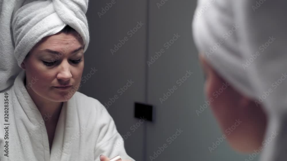 Beautiful girl puts on mascara while looking in the mirror. The camera shows her reflection in the mirror in an over the shoulder frame.
