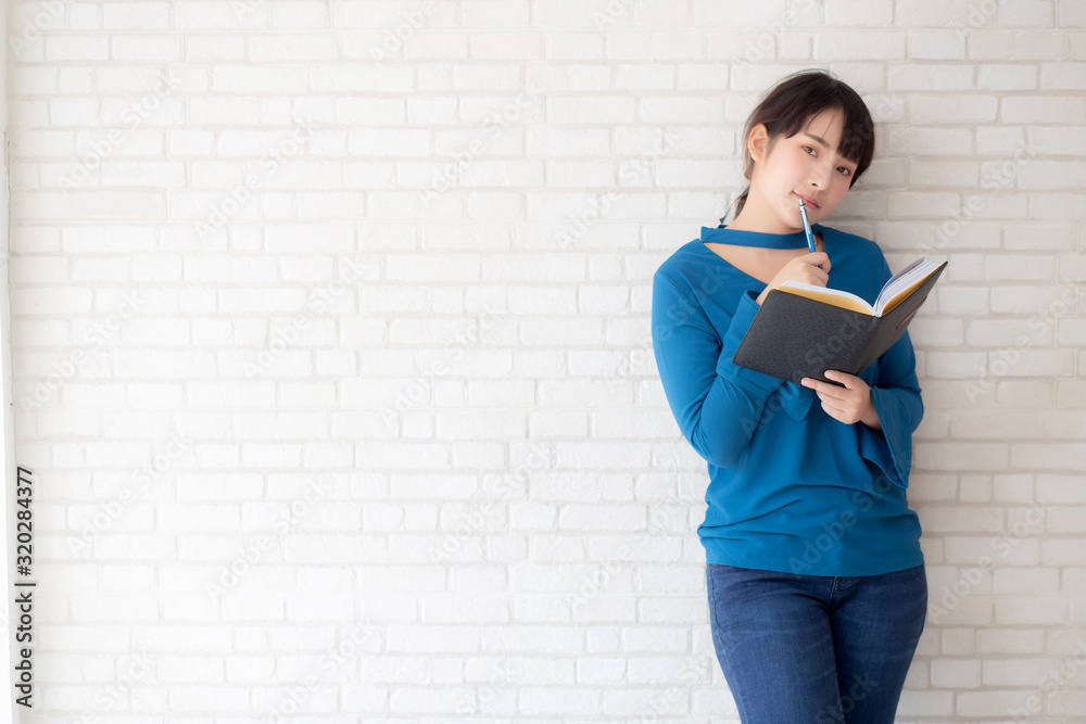 Beautiful asian woman smiling standing thinking and writing notebook on concrete cement white background at home, girl homework on book, education and lifestyle concept. Beautiful asian woman smiling standing thinking and writing notebook on concrete cement white background at home, girl homework on book, education and lifestyle concept.