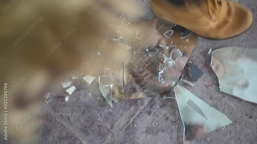 A young woman is smashing pieces of a mirror showing her own reflection with her boots on a dirty, dusty, concrete floor. Angry and furious with energy.
