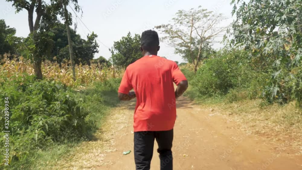 Slow motion shot tracking from behind a young African man jogging on a rural dirt road in Africa during the hot day. Slow motion shot tracking from behind a young African man jogging on a rural dirt road in Africa during the hot day.