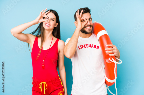 Young lifeguard couple isolated excited keeping ok gesture on eye.