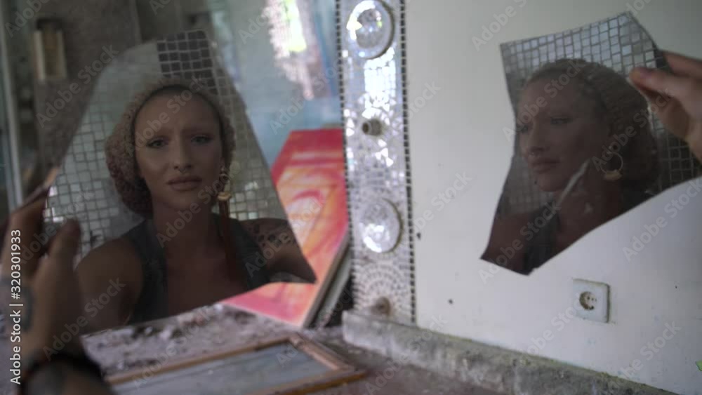 A young woman looks upset and sad into two broken pieces of a mirror that she holds in front of her, sitting by herself in a dark and abandoned room.
