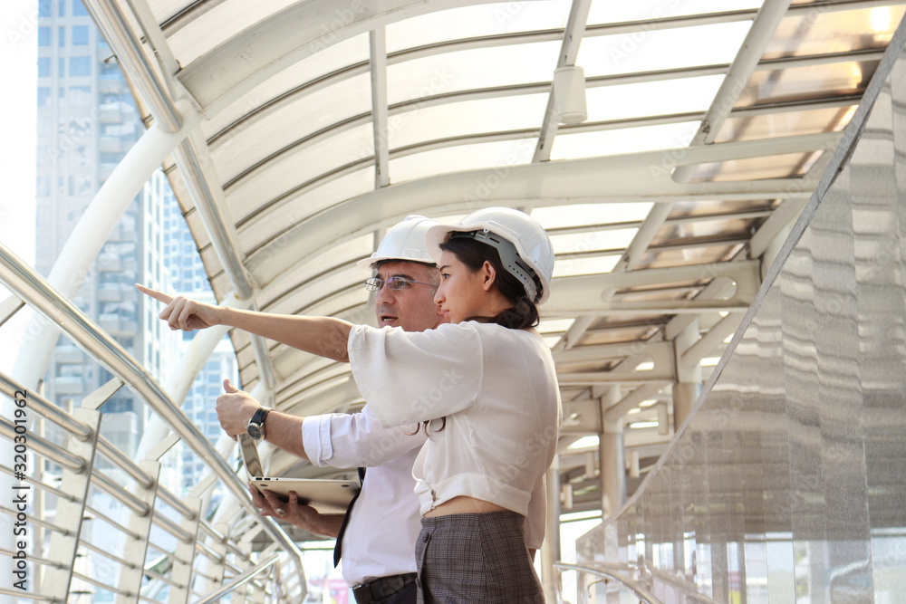 Two engineer wearing white safety helmet using a laptop together, male and female discussing about project work, pointing something, while walking on the way outside office in downtown city