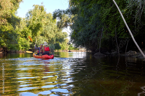 Man and a woman paddle kayak on a river near the shore overgrown with willows at rays of sun at the morning