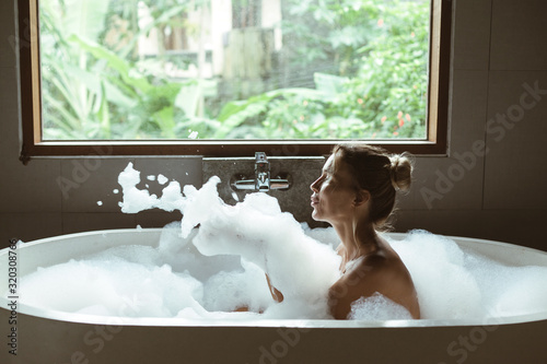 Fototapete Woman relaxing in foam bath with bubbles in dark bathroom by window