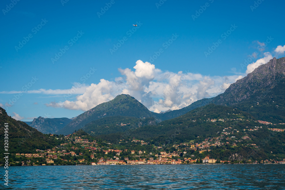 Beautiful summer landscape of Como Lake in Italy, the small town with colorful houses Menaggio, green mountain hills with dense forests, and a small sightseeing seaplane flying in the clear blue sky.