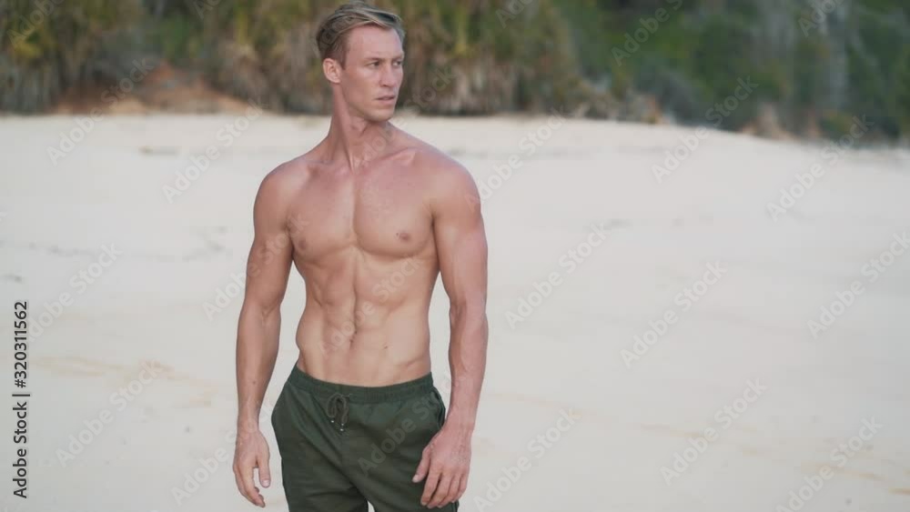 Handsome shirtless man with muscular body stands on beach and looks at ocean