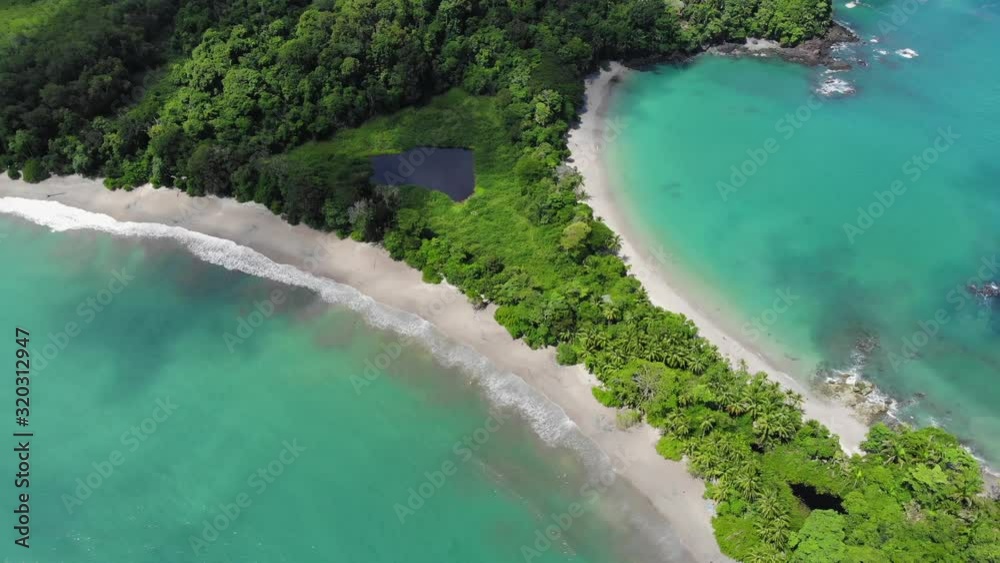 Aerial shot of beach at Cathedral Point, Manuel Antonio National Park, Costa Rica. Sunny weather, clear blue green water, cloud shades over turquoise water. Flying backwards.
