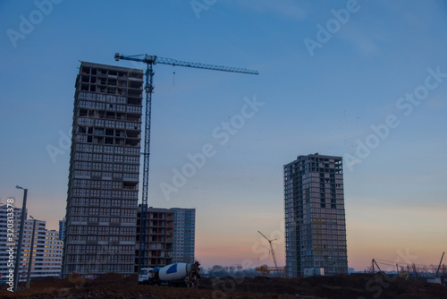 Silhouettes of tower cranes constructing a new residential building at a construction site against sunset background. Heavy mixer concrete truck waiting for to be loaded concrete. Renovation program