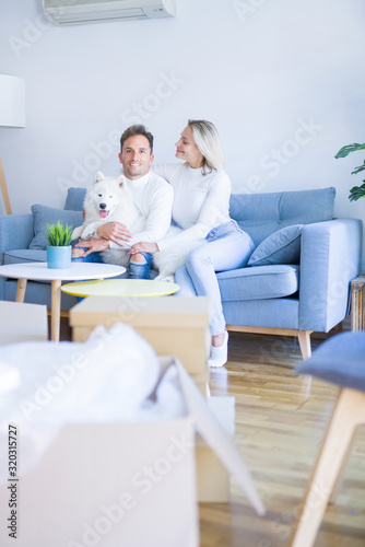 Young beautiful couple with dog sitting on the sofa at new home around cardboard boxes