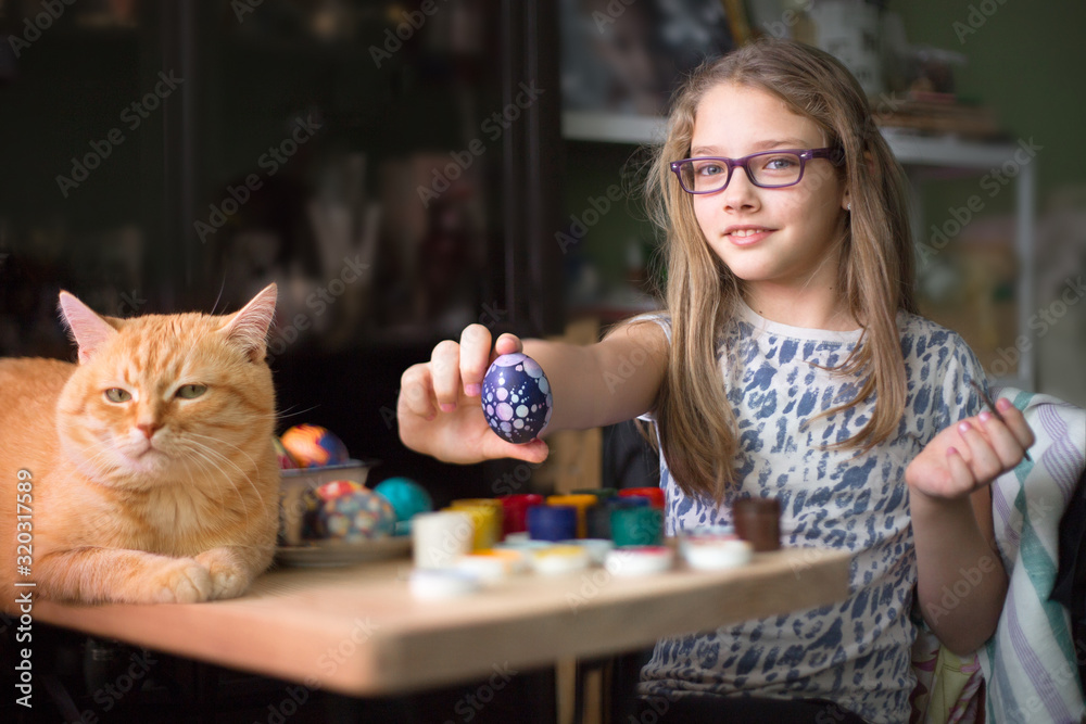 Fototapeta premium Teen girl paints Easter eggs, her ginger cat lies on the table.