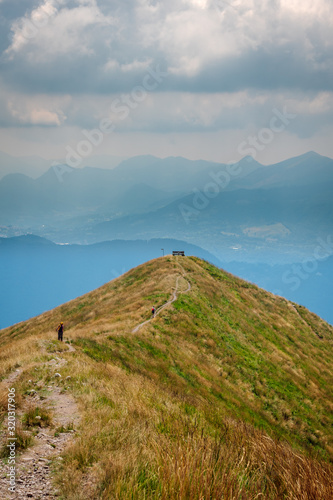 Footpath with two hikers along the ridge of the mountain with a wooden bench at the end with panoramic views over the valley and high mountain ranges in the distance, above Lake Lugano in Swiss Alps.