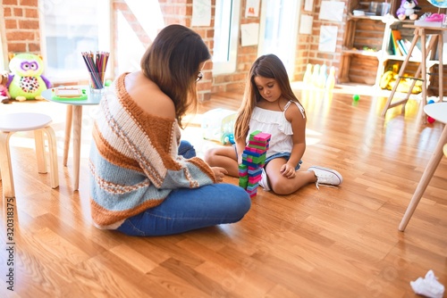 Beautiful teacher and toddler playing with wooden building blocks around lots of toys at kindergarten