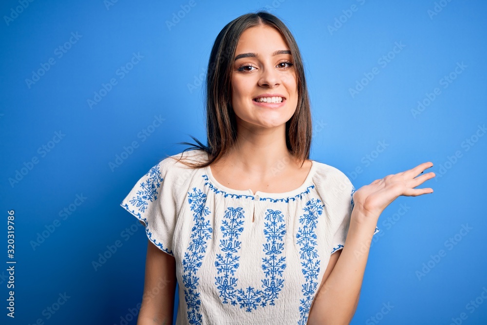Young beautiful brunette woman wearing casual t-shirt standing over blue background smiling cheerful presenting and pointing with palm of hand looking at the camera. Young beautiful brunette woman wearing casual t-shirt standing over blue background smiling cheerful presenting and pointing with palm of hand looking at the camera.