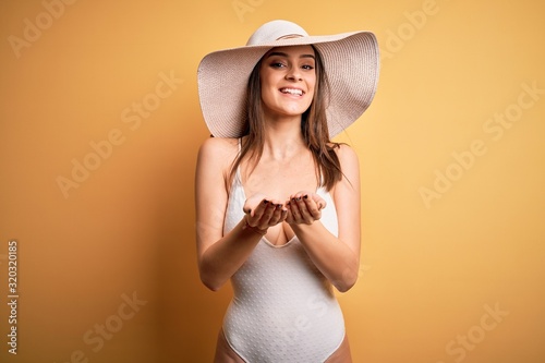 Young beautiful brunette woman on vacation wearing swimsuit and summer hat Smiling with hands palms together receiving or giving gesture. Hold and protection
