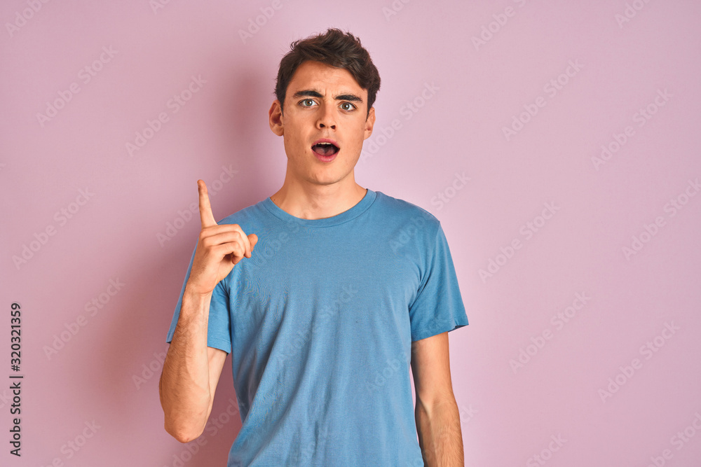 Teenager boy wearing casual t-shirt standing over blue isolated background pointing finger up with successful idea. Exited and happy. Number one. Teenager boy wearing casual t-shirt standing over blue isolated background pointing finger up with successful idea. Exited and happy. Number one.