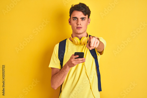 Teenager boy wearing headphones and using smartphone over isolated background pointing with finger to the camera and to you, hand sign, positive and confident gesture from the front
