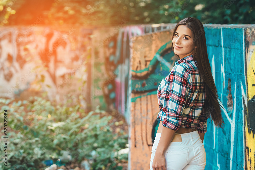 Street photo of beautiful girl. Dressed up in colorful shirt and white jeans. Standing on the background with graffiti. sunshine background