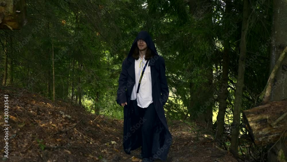 Wandering Missionary Medieval Monk Across Countryside. Man In Black Cloak Coat With Hood Walks On Rural Road In Mountain Forest. Slow Motion Wandering Missionary Medieval Monk Across Countryside. Man In Black Cloak Coat With Hood Walks On Rural Road In Mountain Forest. Slow Motion