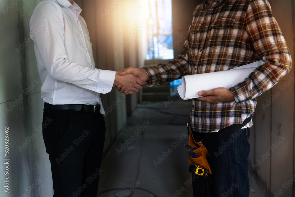Naklejka premium engineer worker holding blueprint shake hand with house agent in construction site