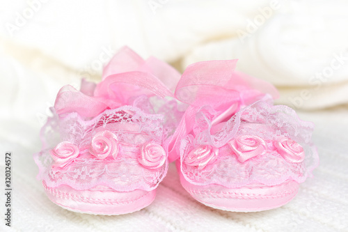 Pink baby booties from the front, on white crochet blanket. Shoes with lace, shiny ribbon and silk rose decoration.