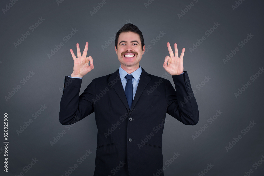 Glad attractive man shows ok sign with both hands as expresses approval, has cheerful expression. Photo of beautiful female has appealing appearance, being optimistic. Standing against gray wall. Glad attractive man shows ok sign with both hands as expresses approval, has cheerful expression. Photo of beautiful female has appealing appearance, being optimistic. Standing against gray wall.