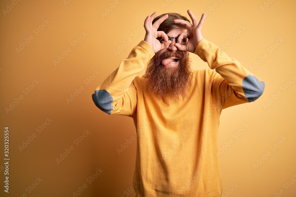 Handsome Irish redhead man with beard wearing glasses over yellow isolated background doing ok gesture like binoculars sticking tongue out, eyes looking through fingers. Crazy expression.